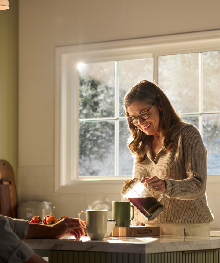 A warmly lit kitchen featuring a large Restorations Series window that frames a soft, sunlit outdoor view. A woman wearing glasses smiles as she pours coffee from a glass carafe into a mug on a marble kitchen island. A man sits across from her, partially visible, with a mug and fresh strawberries nearby. The space includes light cabinetry, a fruit basket on the counter, and gentle morning sunlight streaming through the multi-pane window.