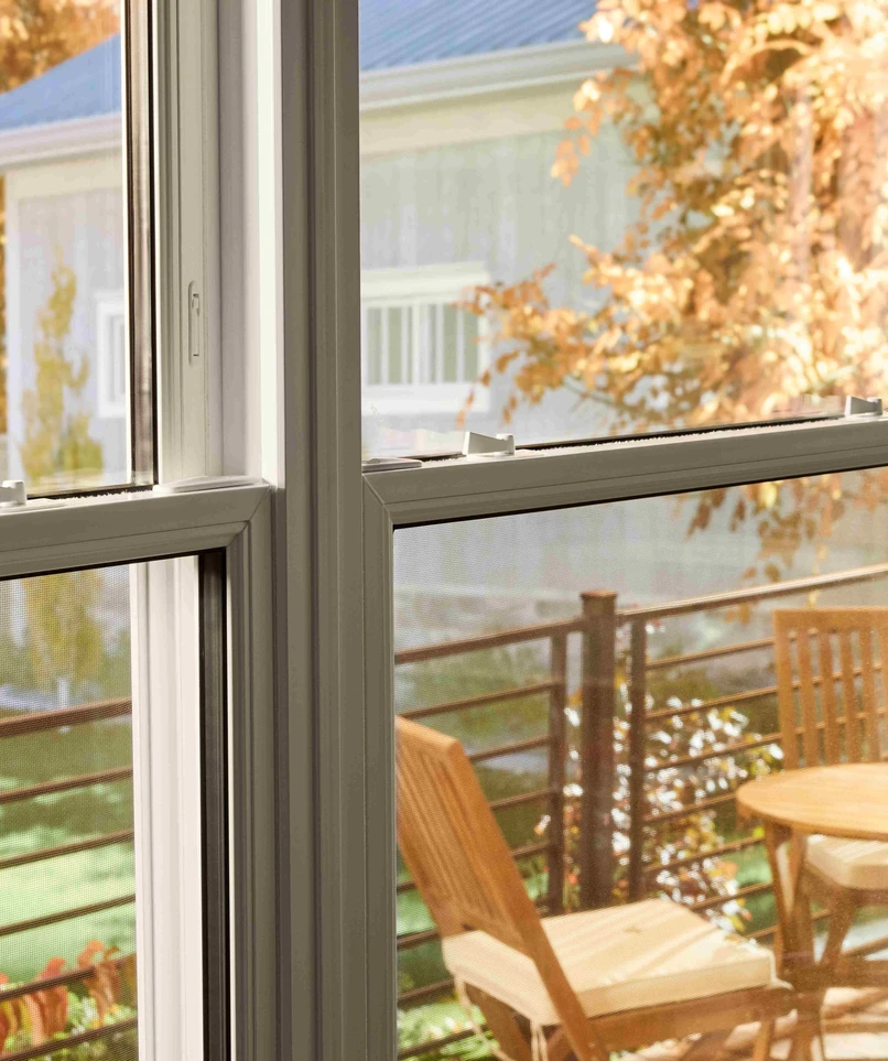View through a double-pane window onto a small outdoor deck with a wooden table and chairs, framed by autumn foliage and neighboring houses in soft daylight.