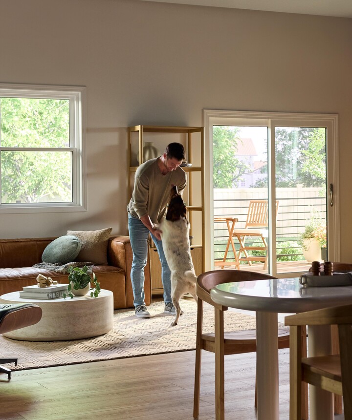 A bright, modern living and dining area featuring large Vanguard Series windows that fill the space with natural light and views of lush greenery outside. A man stands near the center of the room playfully holding his medium-sized dog upright on its hind legs. The room includes a brown leather sofa with accent pillows, a round white coffee table with plants and books, a wooden dining table with chairs, and a sliding glass door leading to a small outdoor patio with additional seating.