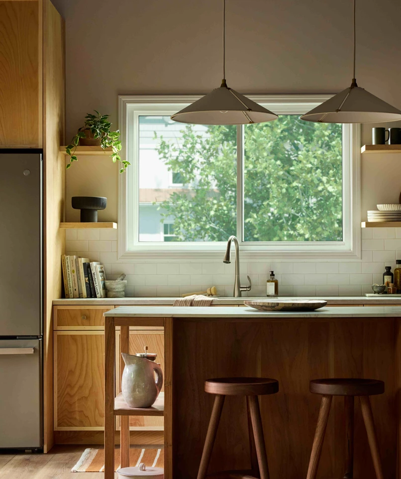 Modern kitchen with warm wood cabinetry and a central island, illuminated by natural light from a large Vanguard Series horizontal slider window above the sink, showing leafy green trees outside.