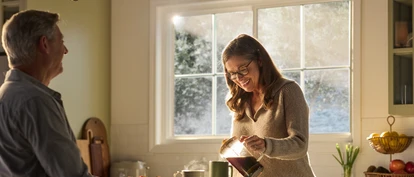 A warmly lit kitchen featuring a large Restorations Series window that frames a soft, sunlit outdoor view. A woman wearing glasses smiles as she pours coffee from a glass carafe into a mug on a marble kitchen island. A man sits across from her, partially visible, with a mug and fresh strawberries nearby. The space includes light cabinetry, a fruit basket on the counter, and gentle morning sunlight streaming through the multi-pane window.