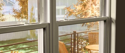 View through a double-pane window onto a small outdoor deck with a wooden table and chairs, framed by autumn foliage and neighboring houses in soft daylight.