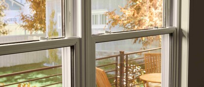 View through a double-pane window onto a small outdoor deck with a wooden table and chairs, framed by autumn foliage and neighboring houses in soft daylight.