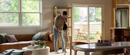 A bright, modern living and dining area featuring large Vanguard Series windows that fill the space with natural light and views of lush greenery outside. A man stands near the center of the room playfully holding his medium-sized dog upright on its hind legs. The room includes a brown leather sofa with accent pillows, a round white coffee table with plants and books, a wooden dining table with chairs, and a sliding glass door leading to a small outdoor patio with additional seating.