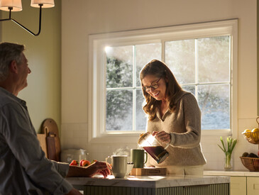 A warmly lit kitchen featuring a large Restorations Series window that frames a soft, sunlit outdoor view. A woman wearing glasses smiles as she pours coffee from a glass carafe into a mug on a marble kitchen island. A man sits across from her, partially visible, with a mug and fresh strawberries nearby. The space includes light cabinetry, a fruit basket on the counter, and gentle morning sunlight streaming through the multi-pane window.