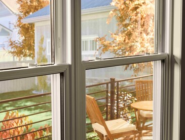 View through a double-pane window onto a small outdoor deck with a wooden table and chairs, framed by autumn foliage and neighboring houses in soft daylight.