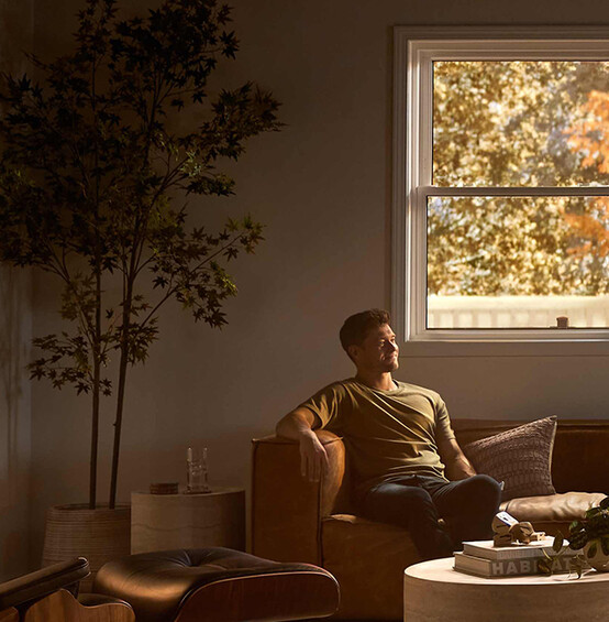 A man relaxes on a leather sofa in a softly lit living room with modern furniture, a round coffee table, and warm autumn light streaming through large windows framed by golden foliage.