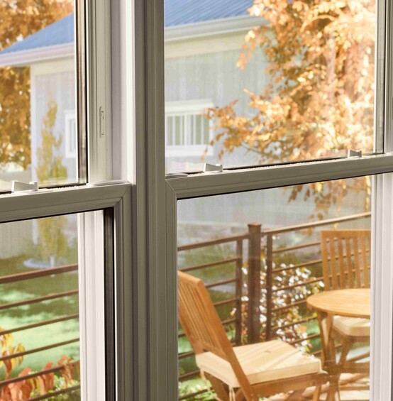 View through a double-pane window onto a small outdoor deck with a wooden table and chairs, framed by autumn foliage and neighboring houses in soft daylight.