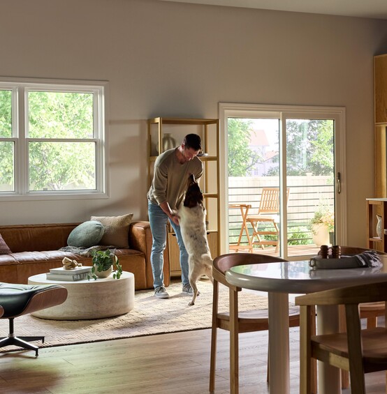 A bright, modern living and dining area featuring large Vanguard Series windows that fill the space with natural light and views of lush greenery outside. A man stands near the center of the room playfully holding his medium-sized dog upright on its hind legs. The room includes a brown leather sofa with accent pillows, a round white coffee table with plants and books, a wooden dining table with chairs, and a sliding glass door leading to a small outdoor patio with additional seating.