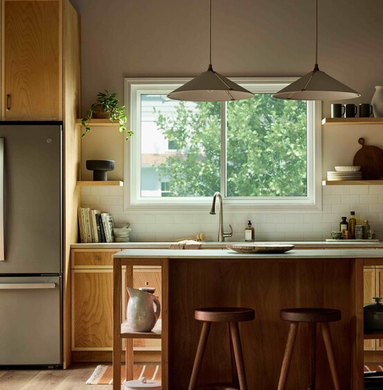 Modern kitchen with warm wood cabinetry and a central island, illuminated by natural light from a large Vanguard Series horizontal slider window above the sink, showing leafy green trees outside.