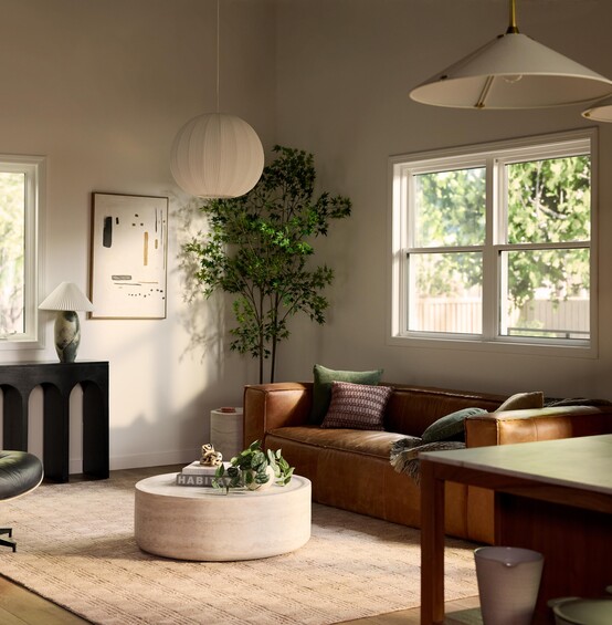 A bright, modern living room with a brown leather sofa, round coffee table, lounge chair, indoor plants, and minimalist decor, lit by natural light from large windows and soft pendant lighting.