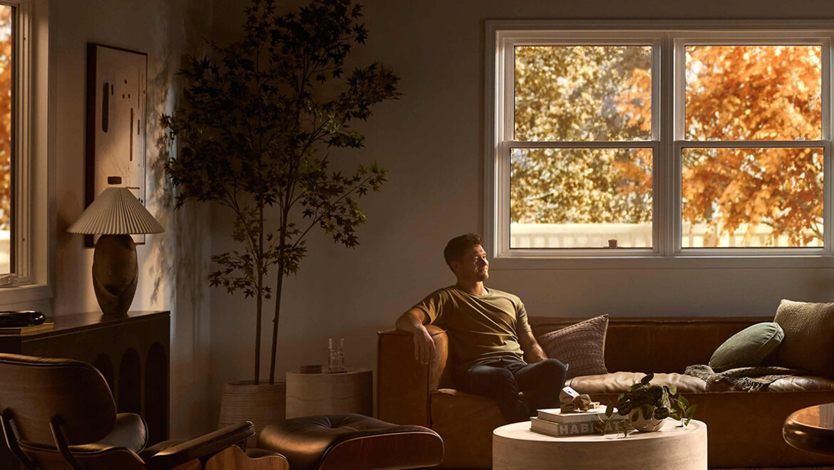 A man relaxes on a leather sofa in a softly lit living room with modern furniture, a round coffee table, and warm autumn light streaming through large windows framed by golden foliage.