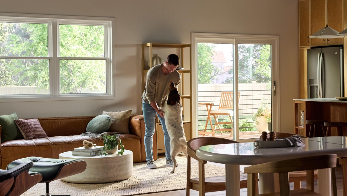 A bright, modern living and dining area featuring large Vanguard Series windows that fill the space with natural light and views of lush greenery outside. A man stands near the center of the room playfully holding his medium-sized dog upright on its hind legs. The room includes a brown leather sofa with accent pillows, a round white coffee table with plants and books, a wooden dining table with chairs, and a sliding glass door leading to a small outdoor patio with additional seating.