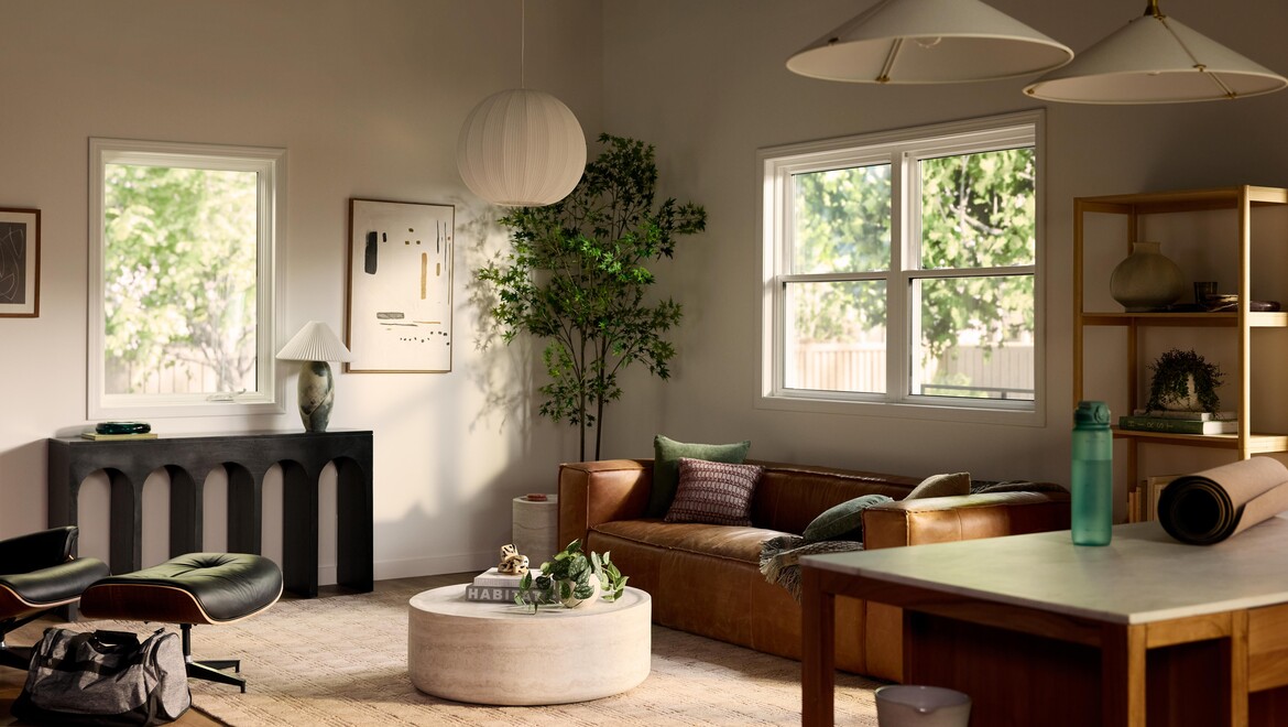 A bright, modern living room with a brown leather sofa, round coffee table, lounge chair, indoor plants, and minimalist decor, lit by natural light from large windows and soft pendant lighting.