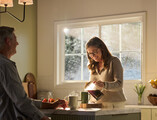 A warmly lit kitchen featuring a large Restorations Series window that frames a soft, sunlit outdoor view. A woman wearing glasses smiles as she pours coffee from a glass carafe into a mug on a marble kitchen island. A man sits across from her, partially visible, with a mug and fresh strawberries nearby. The space includes light cabinetry, a fruit basket on the counter, and gentle morning sunlight streaming through the multi-pane window.
