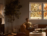 A man relaxes on a leather sofa in a softly lit living room with modern furniture, a round coffee table, and warm autumn light streaming through large windows framed by golden foliage.