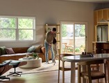 A bright, modern living and dining area featuring large Vanguard Series windows that fill the space with natural light and views of lush greenery outside. A man stands near the center of the room playfully holding his medium-sized dog upright on its hind legs. The room includes a brown leather sofa with accent pillows, a round white coffee table with plants and books, a wooden dining table with chairs, and a sliding glass door leading to a small outdoor patio with additional seating.
