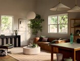 A bright, modern living room with a brown leather sofa, round coffee table, lounge chair, indoor plants, and minimalist decor, lit by natural light from large windows and soft pendant lighting.
