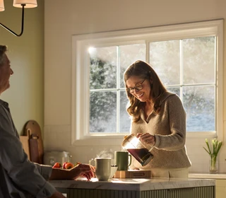 A warmly lit kitchen featuring a large Restorations Series window that frames a soft, sunlit outdoor view. A woman wearing glasses smiles as she pours coffee from a glass carafe into a mug on a marble kitchen island. A man sits across from her, partially visible, with a mug and fresh strawberries nearby. The space includes light cabinetry, a fruit basket on the counter, and gentle morning sunlight streaming through the multi-pane window.