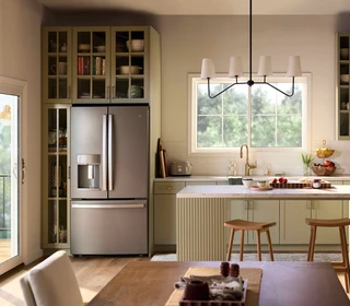 A bright, modern kitchen with sage-green cabinetry, a central island with stools, stainless steel refrigerator, and open shelving, illuminated by natural light from a Restorations Series Horizontal sliding window and a glass door leading to an outdoor deck.