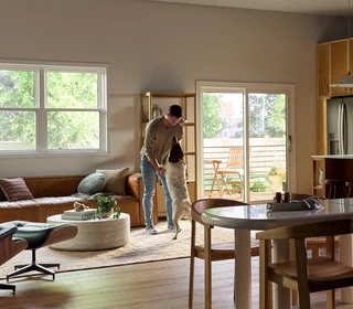 A bright, modern living and dining area featuring large Vanguard Series windows that fill the space with natural light and views of lush greenery outside. A man stands near the center of the room playfully holding his medium-sized dog upright on its hind legs. The room includes a brown leather sofa with accent pillows, a round white coffee table with plants and books, a wooden dining table with chairs, and a sliding glass door leading to a small outdoor patio with additional seating.