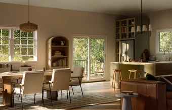 Open-concept living space with a dining area featuring a wooden table and upholstered chairs, a sage green kitchen with island seating and stainless steel refrigerator, brown leather sofa in the living area, and natural light streaming in through multiple white-framed double-hung windows and a sliding glass patio door overlooking trees.