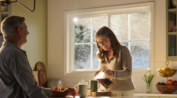 A warmly lit kitchen featuring a large Restorations Series window that frames a soft, sunlit outdoor view. A woman wearing glasses smiles as she pours coffee from a glass carafe into a mug on a marble kitchen island. A man sits across from her, partially visible, with a mug and fresh strawberries nearby. The space includes light cabinetry, a fruit basket on the counter, and gentle morning sunlight streaming through the multi-pane window.