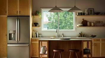 Modern kitchen with warm wood cabinetry and a central island, illuminated by natural light from a large Vanguard Series horizontal slider window above the sink, showing leafy green trees outside.