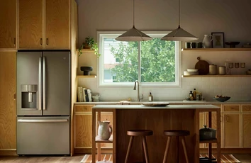 Modern kitchen with warm wood cabinetry and a central island, illuminated by natural light from a large Vanguard Series horizontal slider window above the sink, showing leafy green trees outside.