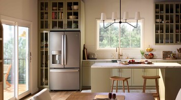 A bright, modern kitchen with sage-green cabinetry, a central island with stools, stainless steel refrigerator, and open shelving, illuminated by natural light from a Restorations Series Horizontal sliding window and a glass door leading to an outdoor deck.