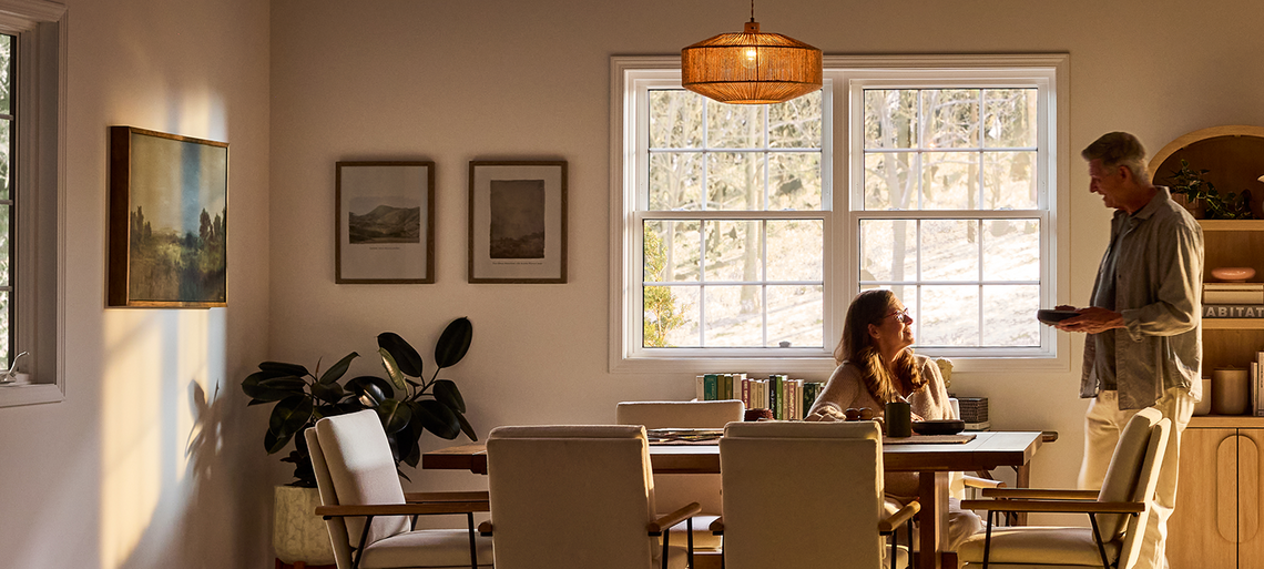 A sunlit dining room with Restorations Series windows, where an older couple shares breakfast at a wooden table, surrounded by soft neutral furnishings, framed artwork, indoor plants, and warm natural light filtering through large grid-pane windows overlooking trees.