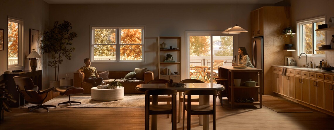 A wide view of an open-plan living room and kitchen where a man relaxes on a sofa and a woman sits at a kitchen island, with warm wood cabinetry, modern furniture, pendant lights, and large windows showing autumn trees and an outdoor deck.