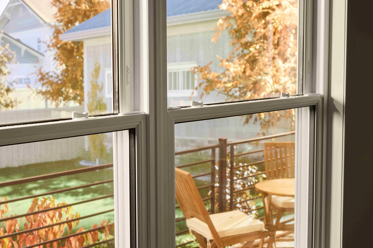 View through a double-pane window onto a small outdoor deck with a wooden table and chairs, framed by autumn foliage and neighboring houses in soft daylight.