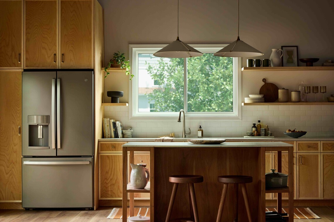 Modern kitchen with warm wood cabinetry and a central island, illuminated by natural light from a large Vanguard Series horizontal slider window above the sink, showing leafy green trees outside.