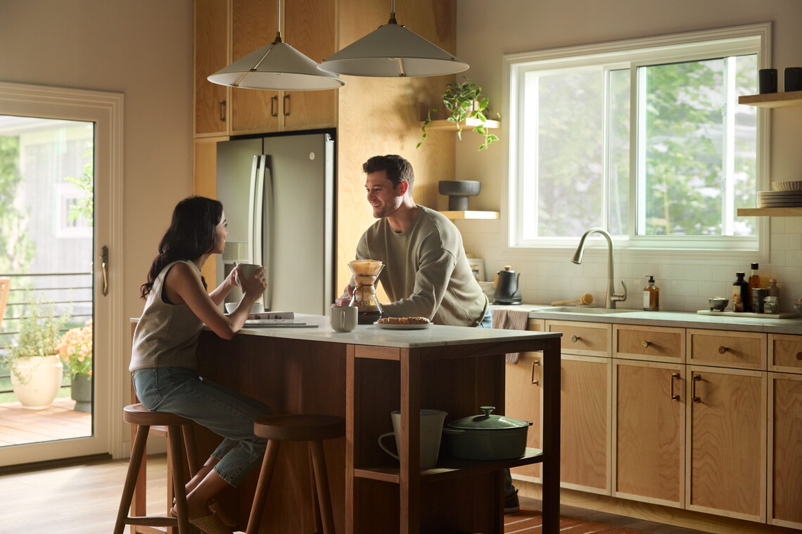 A man and a woman chat over coffee at a kitchen island in a bright, modern kitchen with light wood cabinets, pendant lights, and a large Vanguard Series horizontal slider window letting in natural daylight.