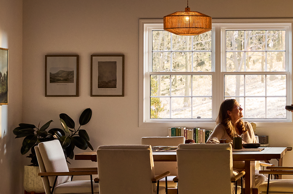 A sunlit dining room with Restorations Series windows, where an older couple shares breakfast at a wooden table, surrounded by soft neutral furnishings, framed artwork, indoor plants, and warm natural light filtering through large grid-pane windows overlooking trees.