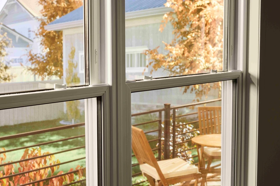 View through a double-pane window onto a small outdoor deck with a wooden table and chairs, framed by autumn foliage and neighboring houses in soft daylight.