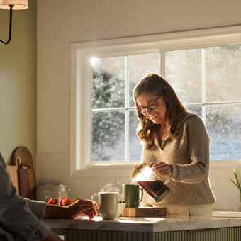 A warmly lit kitchen featuring a large Restorations Series window that frames a soft, sunlit outdoor view. A woman wearing glasses smiles as she pours coffee from a glass carafe into a mug on a marble kitchen island. A man sits across from her, partially visible, with a mug and fresh strawberries nearby. The space includes light cabinetry, a fruit basket on the counter, and gentle morning sunlight streaming through the multi-pane window.