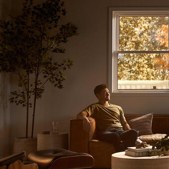 A man relaxes on a leather sofa in a softly lit living room with modern furniture, a round coffee table, and warm autumn light streaming through large windows framed by golden foliage.