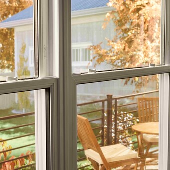 View through a double-pane window onto a small outdoor deck with a wooden table and chairs, framed by autumn foliage and neighboring houses in soft daylight.