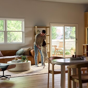 A bright, modern living and dining area featuring large Vanguard Series windows that fill the space with natural light and views of lush greenery outside. A man stands near the center of the room playfully holding his medium-sized dog upright on its hind legs. The room includes a brown leather sofa with accent pillows, a round white coffee table with plants and books, a wooden dining table with chairs, and a sliding glass door leading to a small outdoor patio with additional seating.