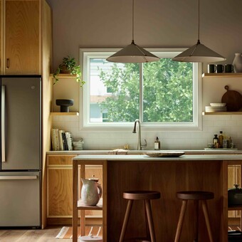 Modern kitchen with warm wood cabinetry and a central island, illuminated by natural light from a large Vanguard Series horizontal slider window above the sink, showing leafy green trees outside.