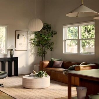 A bright, modern living room with a brown leather sofa, round coffee table, lounge chair, indoor plants, and minimalist decor, lit by natural light from large windows and soft pendant lighting.