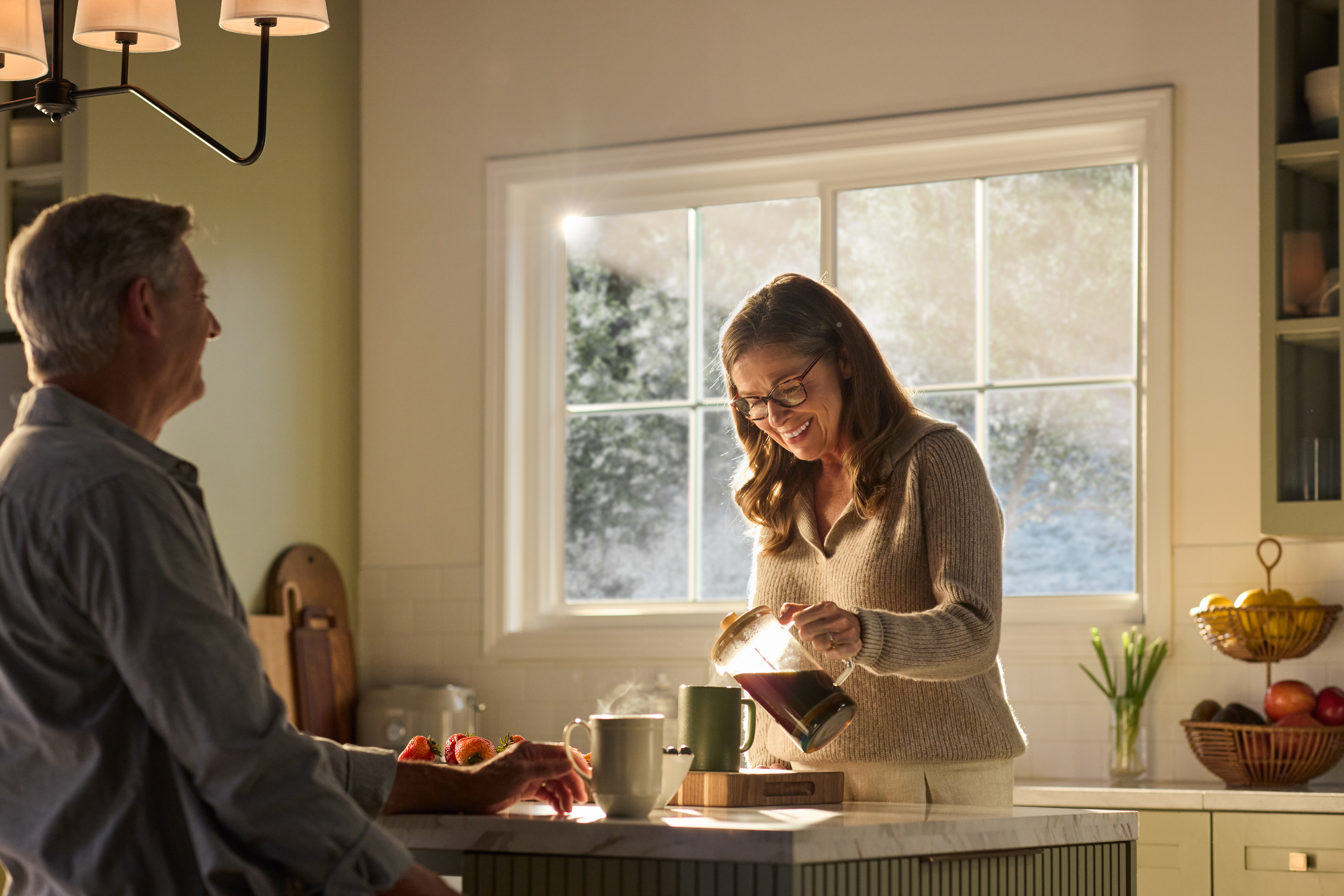 A warmly lit kitchen featuring a large Restorations Series window that frames a soft, sunlit outdoor view. A woman wearing glasses smiles as she pours coffee from a glass carafe into a mug on a marble kitchen island. A man sits across from her, partially visible, with a mug and fresh strawberries nearby. The space includes light cabinetry, a fruit basket on the counter, and gentle morning sunlight streaming through the multi-pane window.