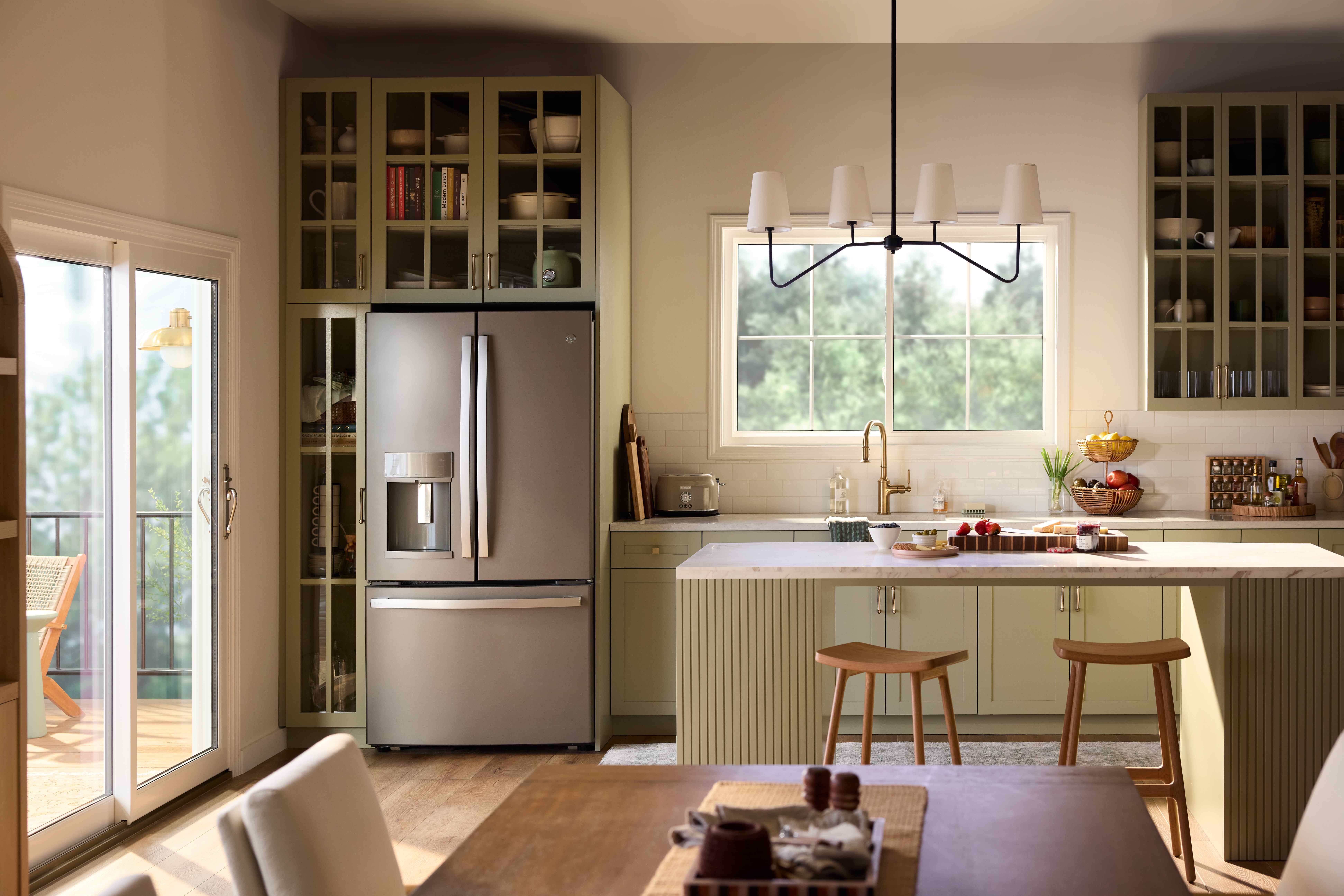 A bright, modern kitchen with sage-green cabinetry, a central island with stools, stainless steel refrigerator, and open shelving, illuminated by natural light from a Restorations Series Horizontal sliding window and a glass door leading to an outdoor deck.