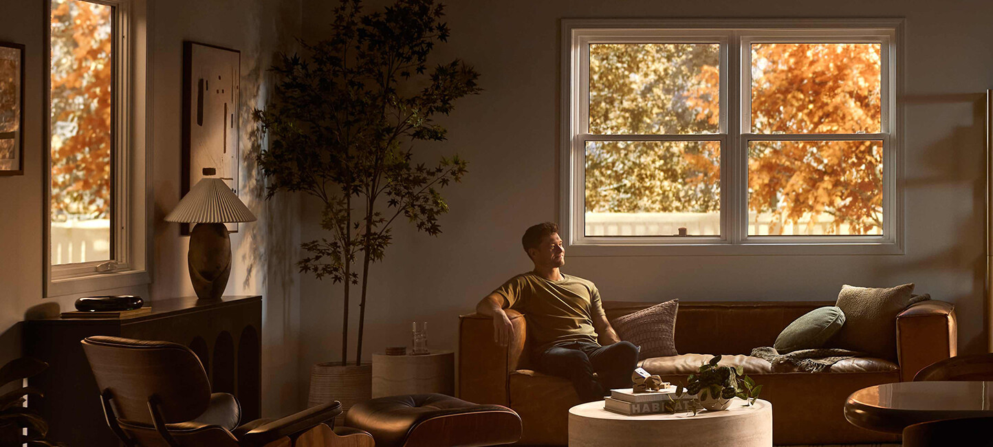 A man relaxes on a leather sofa in a softly lit living room with modern furniture, a round coffee table, and warm autumn light streaming through large windows framed by golden foliage.