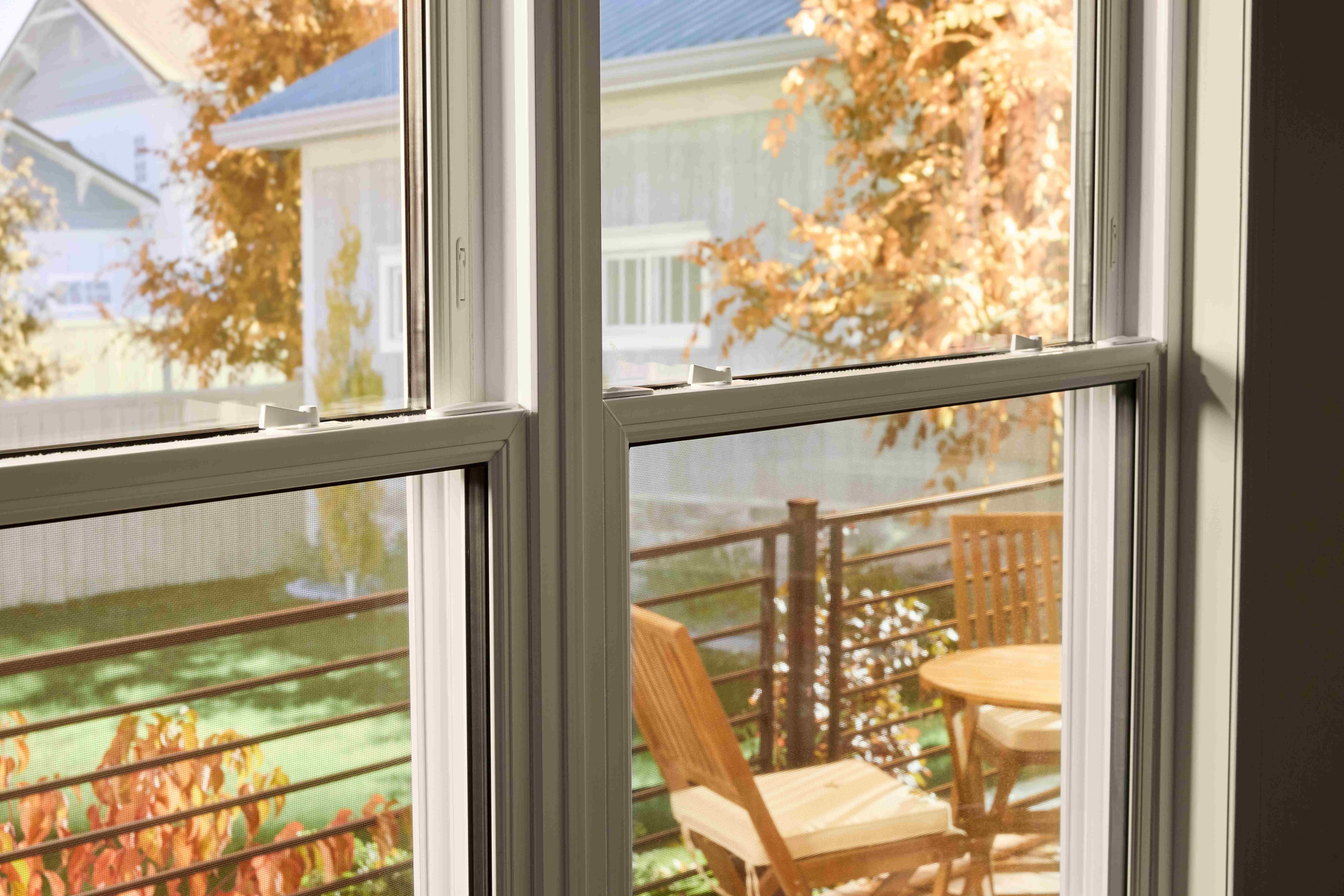 View through a double-pane window onto a small outdoor deck with a wooden table and chairs, framed by autumn foliage and neighboring houses in soft daylight.