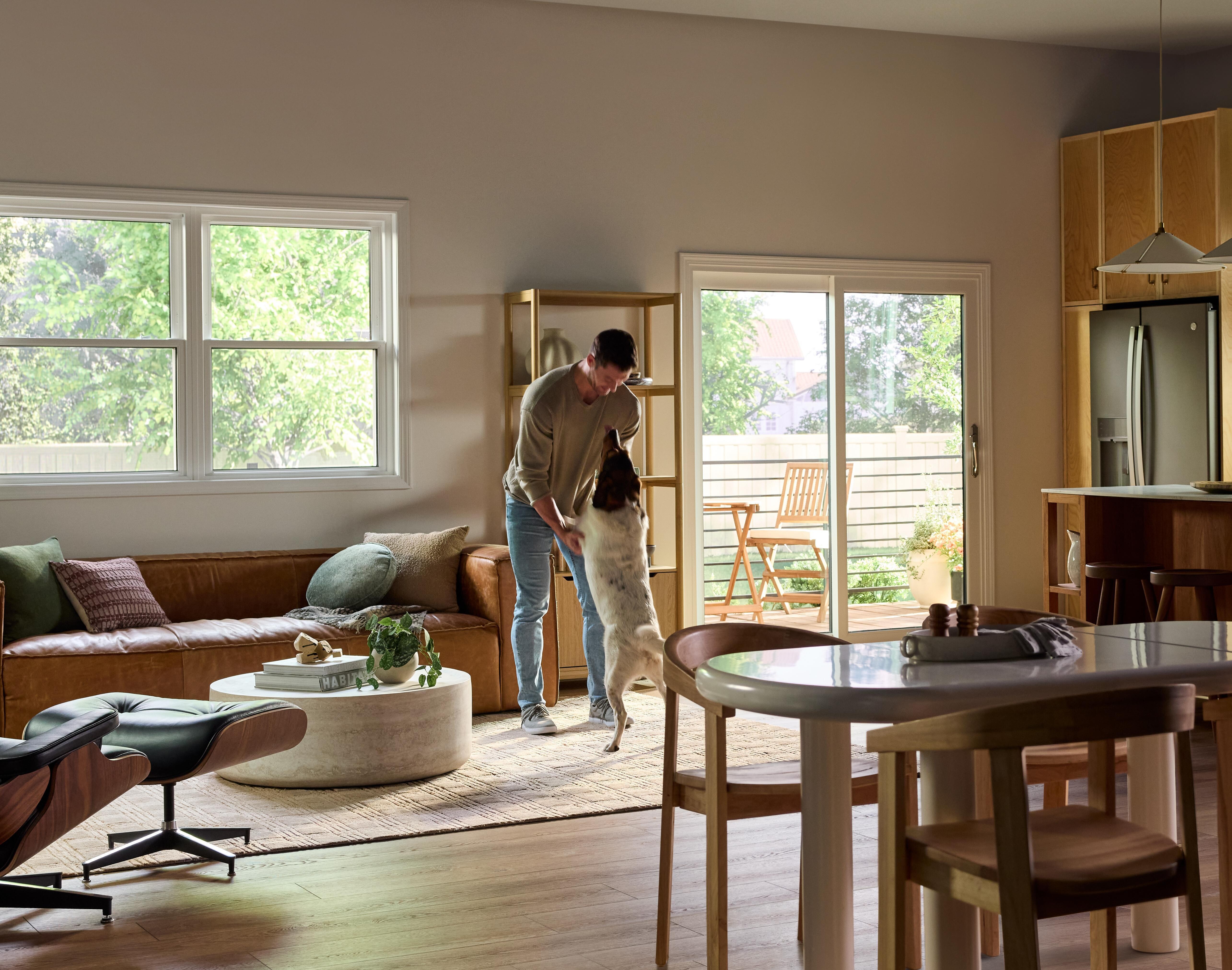 A bright, modern living and dining area featuring large Vanguard Series windows that fill the space with natural light and views of lush greenery outside. A man stands near the center of the room playfully holding his medium-sized dog upright on its hind legs. The room includes a brown leather sofa with accent pillows, a round white coffee table with plants and books, a wooden dining table with chairs, and a sliding glass door leading to a small outdoor patio with additional seating.