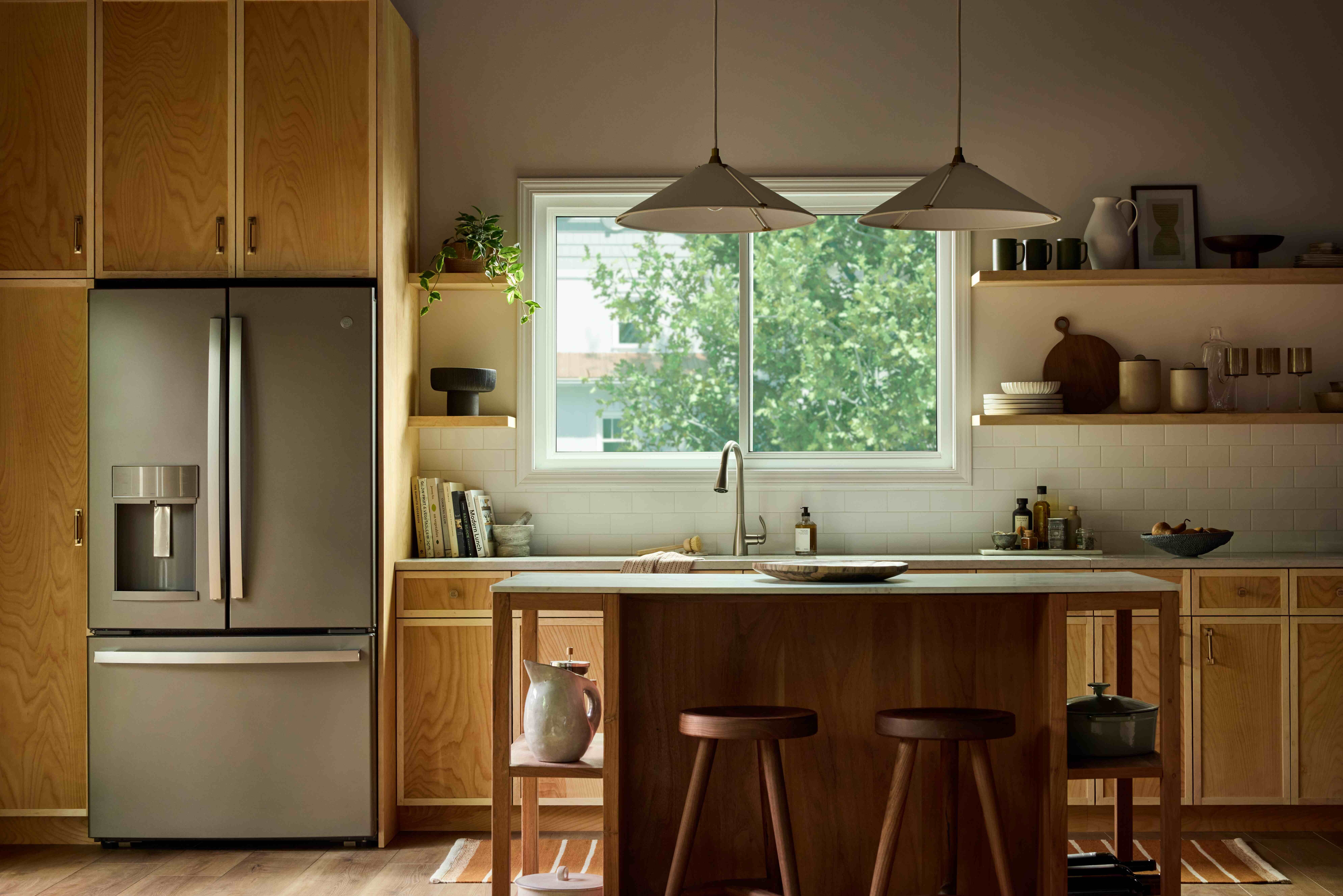 Modern kitchen with warm wood cabinetry and a central island, illuminated by natural light from a large Vanguard Series horizontal slider window above the sink, showing leafy green trees outside.