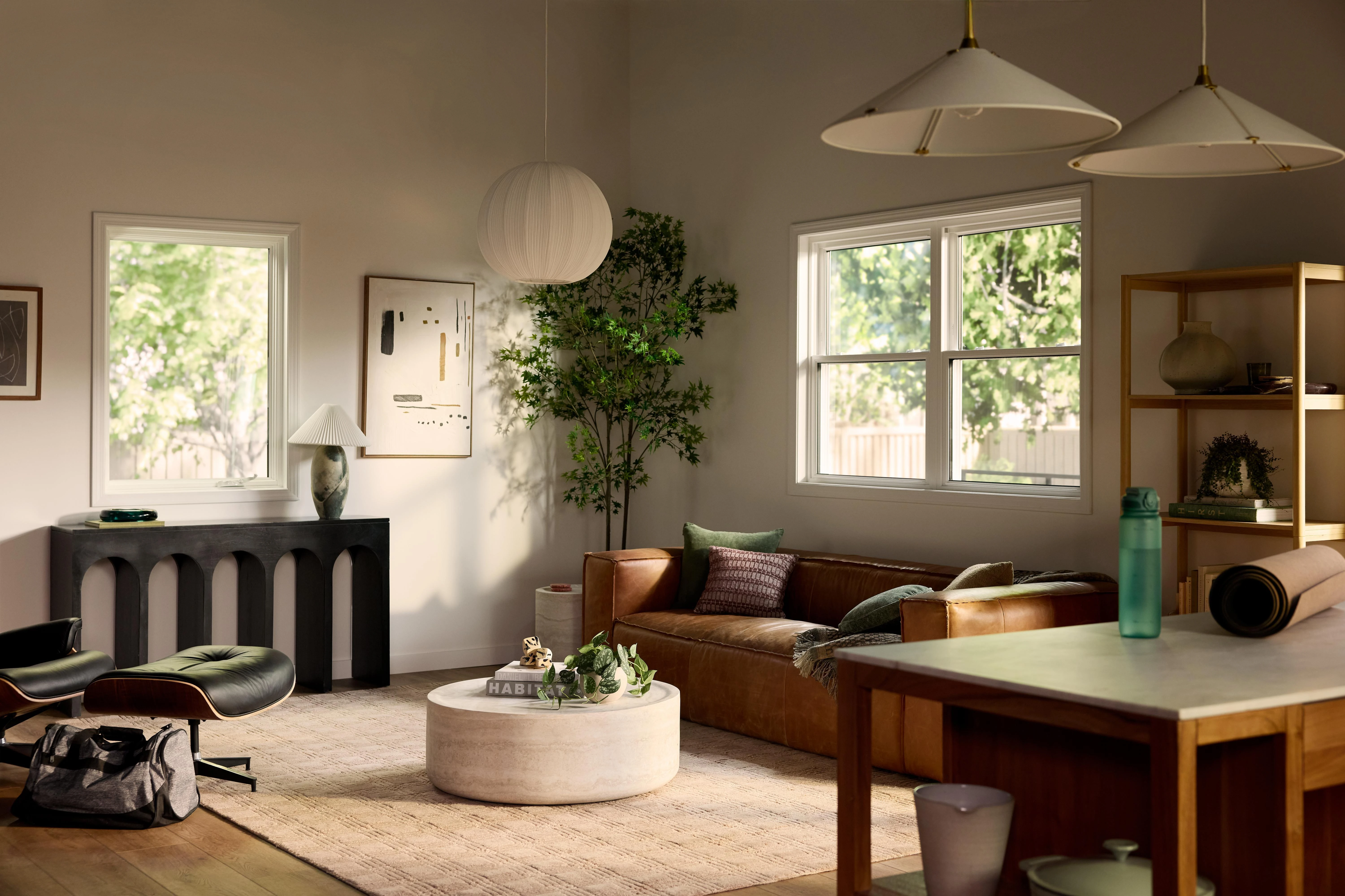 A bright, modern living room with a brown leather sofa, round coffee table, lounge chair, indoor plants, and minimalist decor, lit by natural light from large windows and soft pendant lighting.